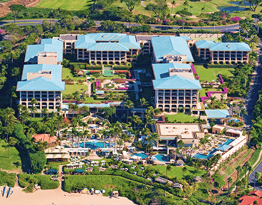 An aerial view of a large coastal resort with blue roofs and multiple swimming pools near the beach.