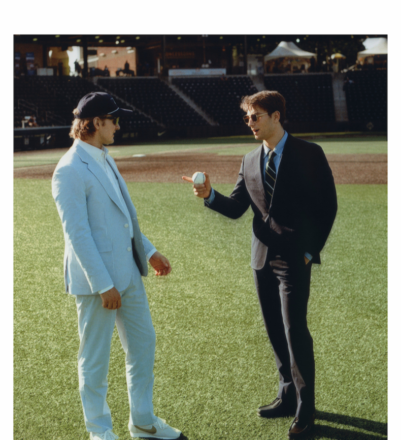 Two men stand on a baseball field in conversation. One wears a fitted navy seersucker suit and striped tie, the other wears a seersucker suit with white sneakers and a baseball cap.