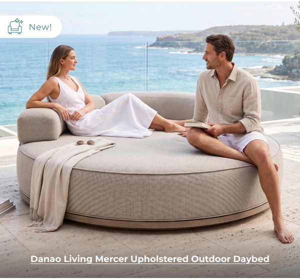 A man and a woman converse while relaxing on a large, round upholstered outdoor daybed situated on a terrace with an ocean backdrop. Image Text: New! Danao Living Mercer Upholstered Outdoor Daybed