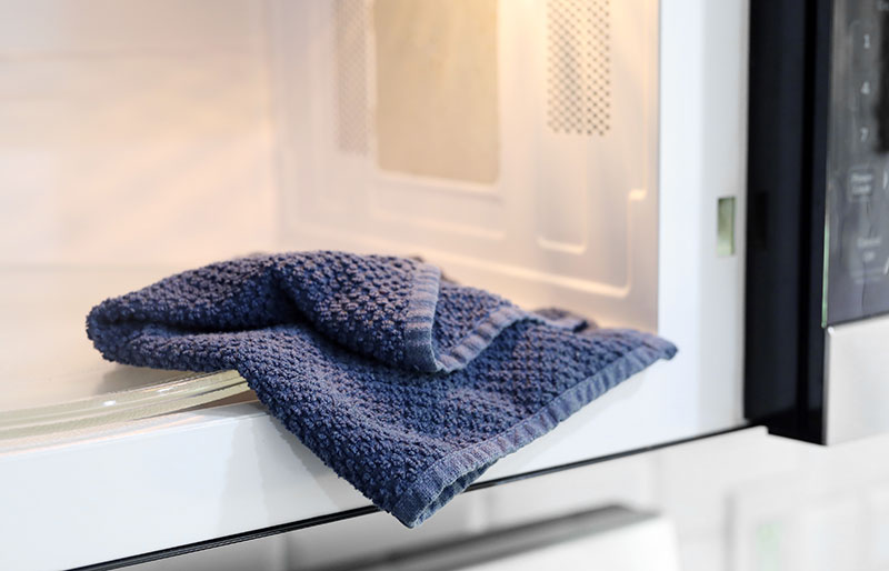 A navy blue towel rests on the open door of a clean microwave; a simple routine for keeping your microwave spotless and Anyday dishes cooking efficiently.