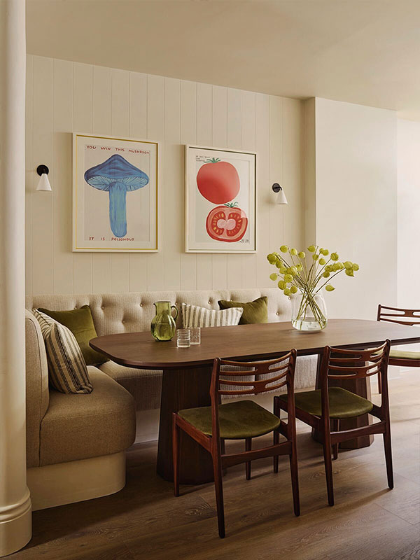 A warm dining space with a wooden table, cushioned bench, and green‑seated chairs. Framed artwork of a blue mushroom and red tomato adds playful character.