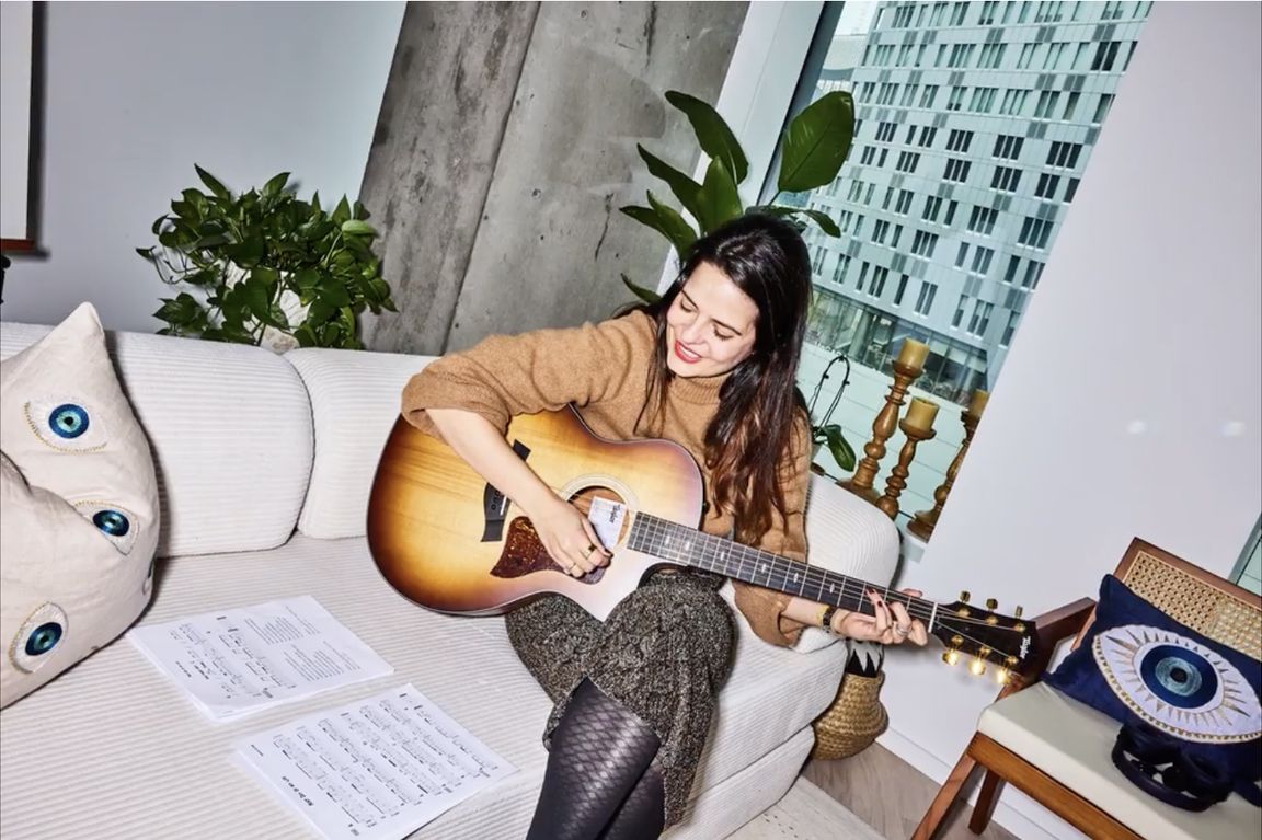 Woman sitting on couch playing acoustic guitar while looking at sheet music