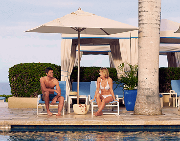 A man and woman relaxing by a pool, sitting on lounge chairs under a large umbrella, with the ocean in the background.