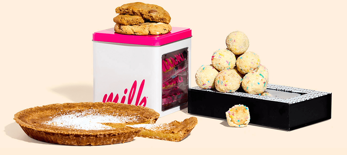 Various Milk Bar desserts, including a Milk Bar Pie, a white and pink cookie tin with unwrapped cookies sitting atop it, and a black box with unwrapped Birthday Truffles stacked upon it. 
