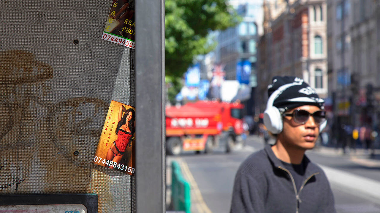 People passing a public telephone box which has sex cards tacked up inside 