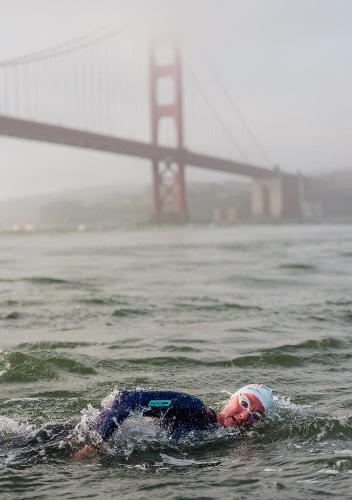A woman in a white bathing cap and a black wet suit swimming near the Golden Gate Bridge.