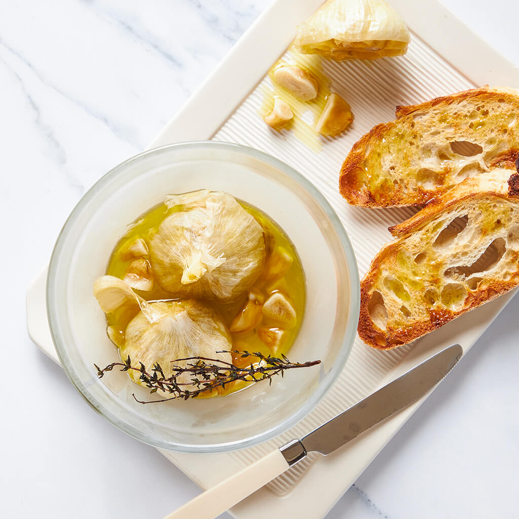 A glass Anyday dish filled with roasted garlic in olive oil, garnished with thyme, alongside toasted bread slices on a white cutting board. This easy microwave-roasted garlic recipe creates soft, caramelized garlic perfect for spreading or adding to dishes.