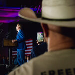 Ken Paxton, wearing a blue suit, stands at a lectern, with a large American flag hanging to his right.