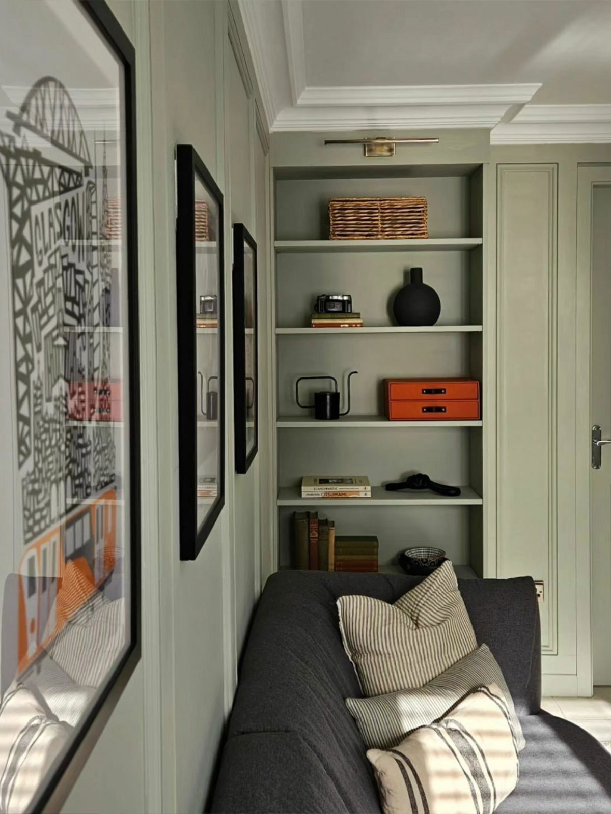 A plump grey sofa invites us to stop and relax in this pale olive green space. Put your feet up and just enjoy the peaceful uncluttered environment. The shelves contain a curated collection of fascinating items: vintage books, a deep grey vase, and an interestingly shaped black watering can, complemented by the orange mini drawer unit.