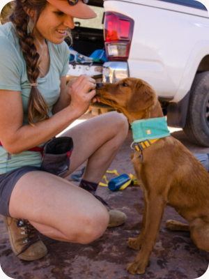A woman with her dog outside giving the dog a treat after putting on their cooling neck gaiter from Ruffwear.
