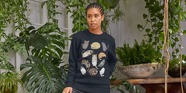 Lady wearing an RSPCA Teemill jumper with prints of cats on. She is stood indoors surrounded by green plants