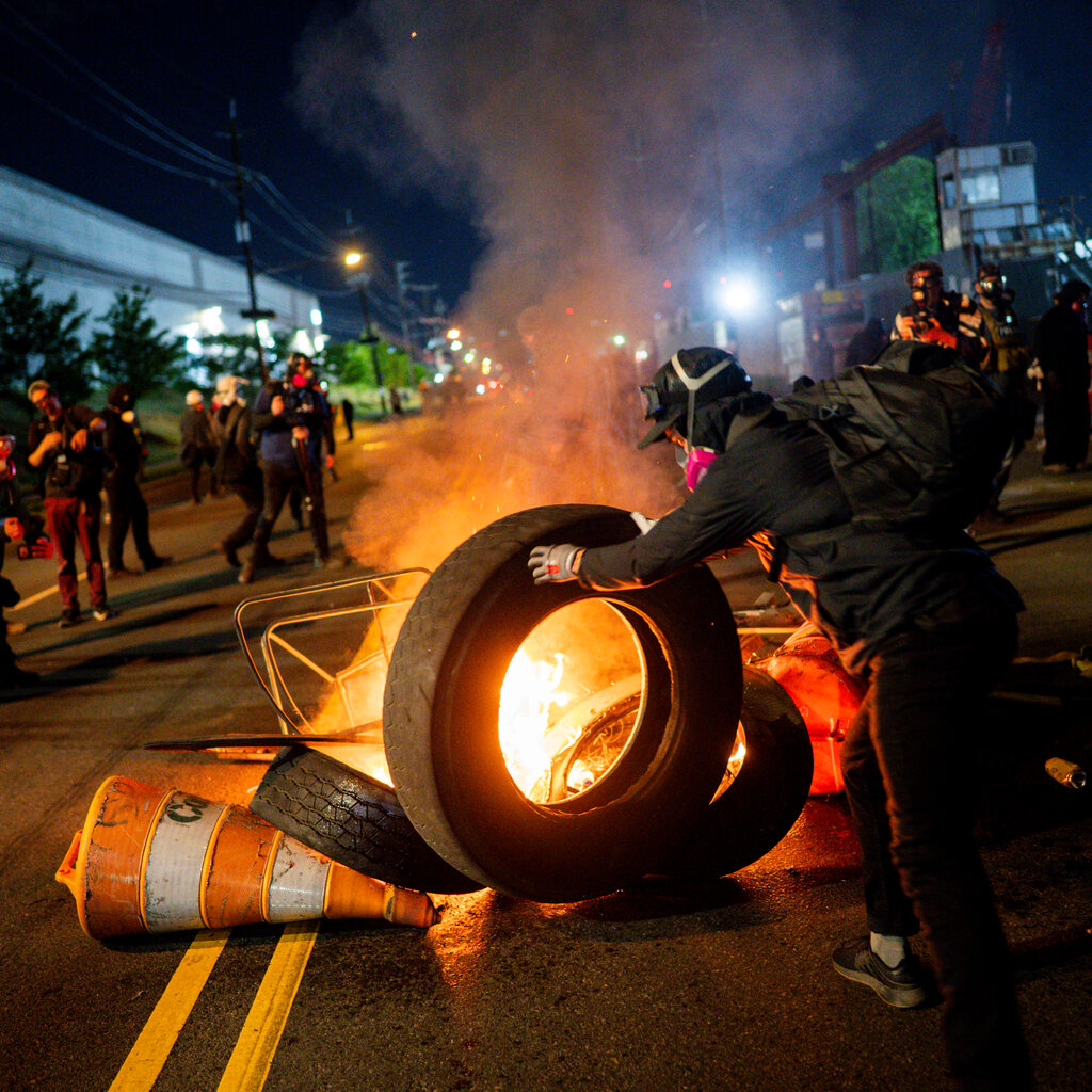 A person in a helmet and a pink-filtered respirator pushes a burning tire on a street at night. Bright orange flames and smoke rise from the tire, with other individuals nearby.