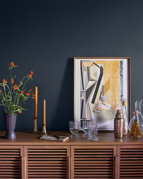 A wood buffet table topped with candles, plants, and other décor, sits in front of a navy blue painted wall.