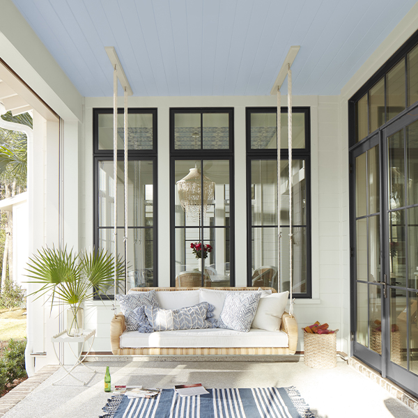 A white porch with window trim painted black, a white floor, blue and white area rug, and a hanging swing.