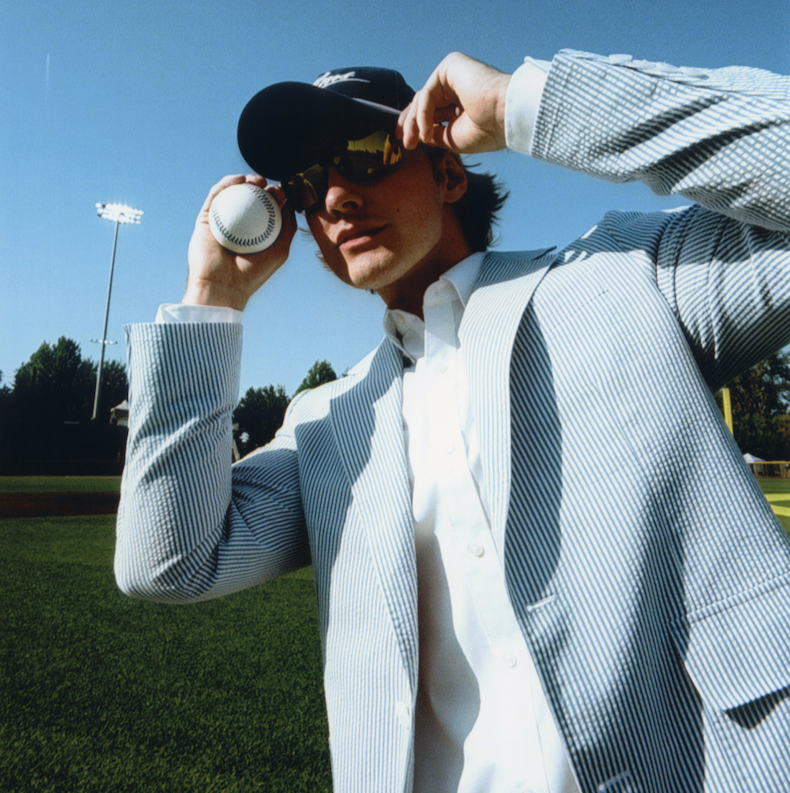 A man stands on a baseball field in bright daylight, holding a baseball next to his face. He wears a pale blue and white seersucker suit, a white shirt, a navy cap, and sunglasses.