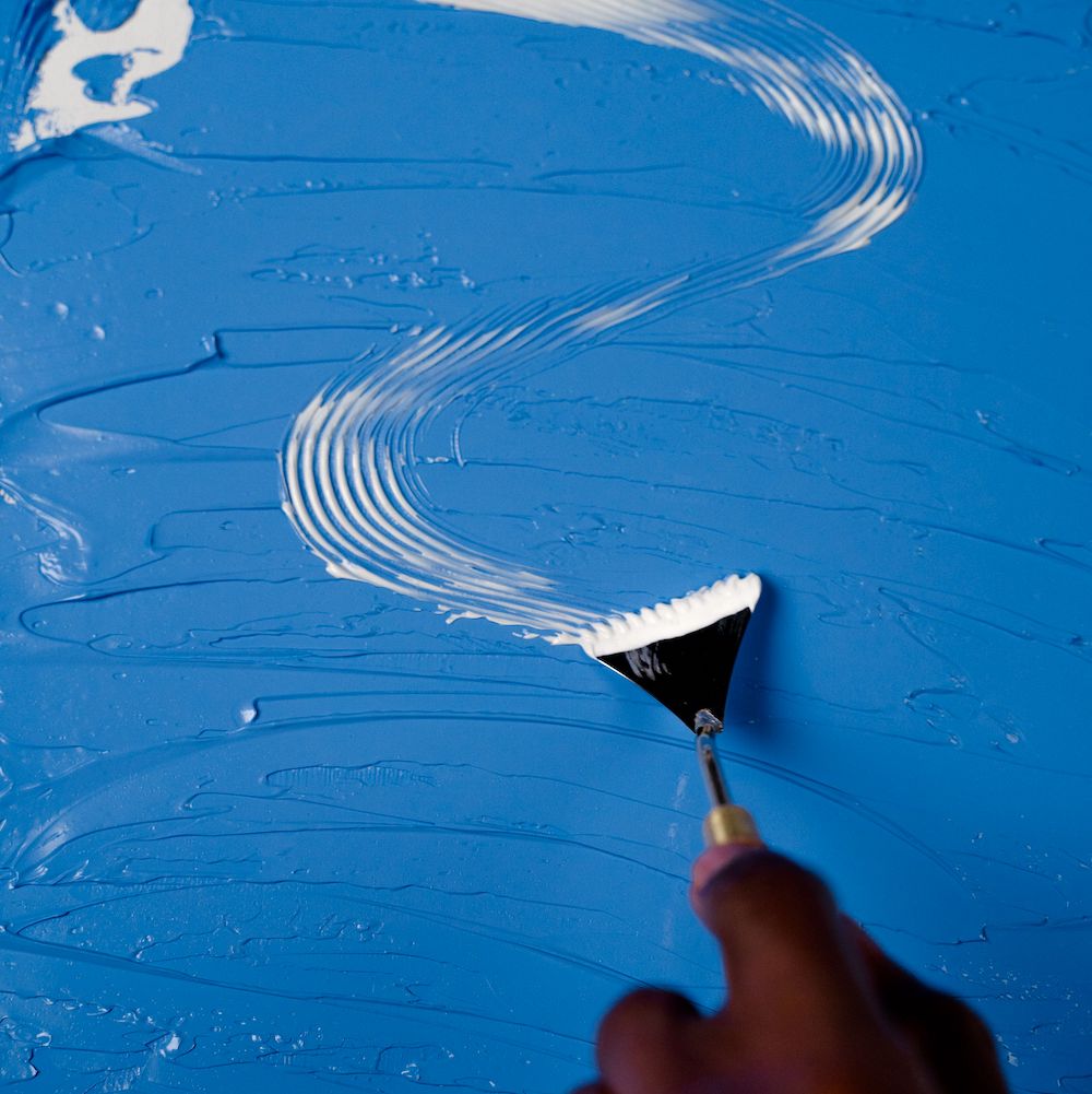 A hand holding a paint spatula spreads a wavy white line over a blue surface.