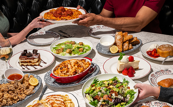 People sharing a restaurant meal at a table filled with several Maggiano’s entrées, including spaghetti and meatballs, salads, lasagna, fried appetizers, desserts, and drinks.