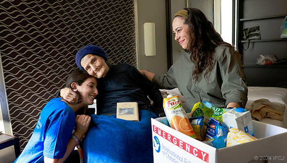 Yael Eckstein and an IFCJ staff member sitting with an elderly woman, next to an IFCJ food box.