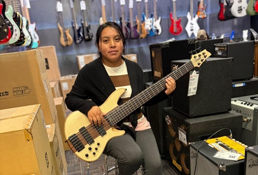 Andrea Pedro-Francisco sitting on a stool and playing a bass, surrounded by amps, boxes, and other instruments on display along the walls