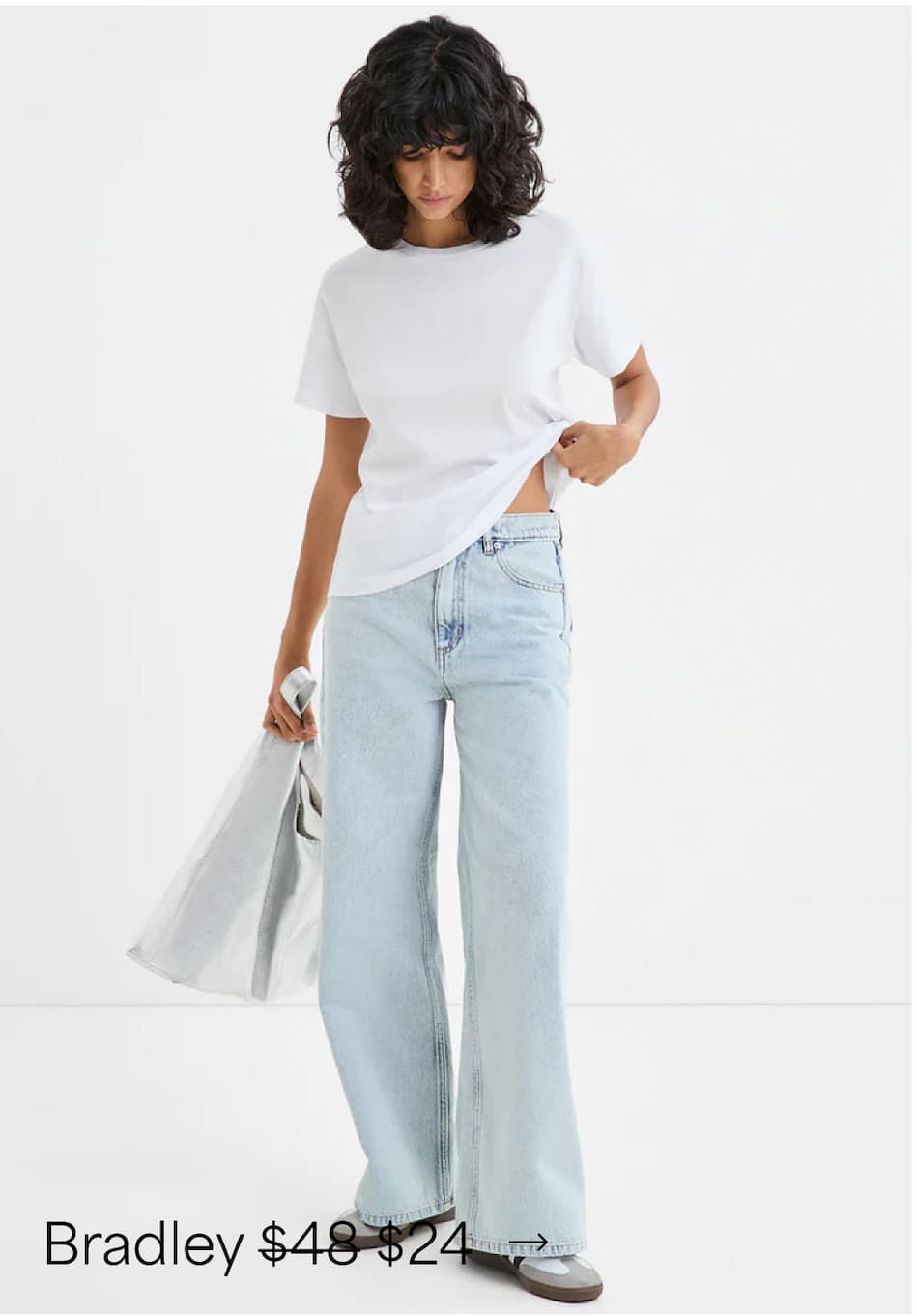 Emma in The Bradley Tee and light-wash wide-leg jeans holds a silver tote against a white backdrop.