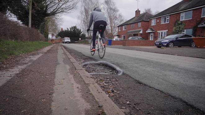 A road rider travels away from the camera with a pothole visible in the foreground on a residential street.