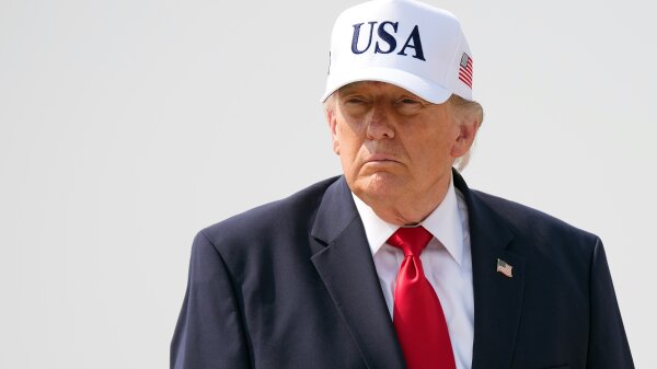 President Trump, from the shoulders up, wearing a suit with a red tie. He wears a white cap with "USA" in dark blue written across it.