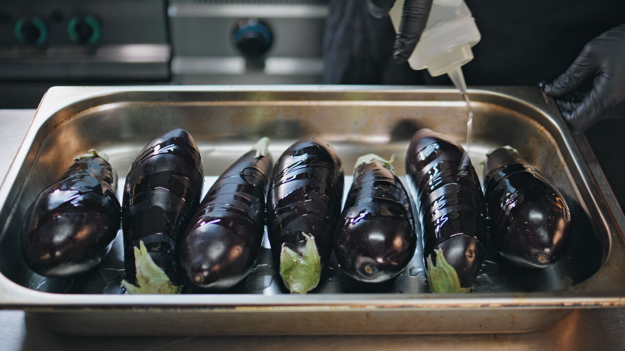 Hand pouring oil over eggplants in metal tray restaurant kitchen closeup.