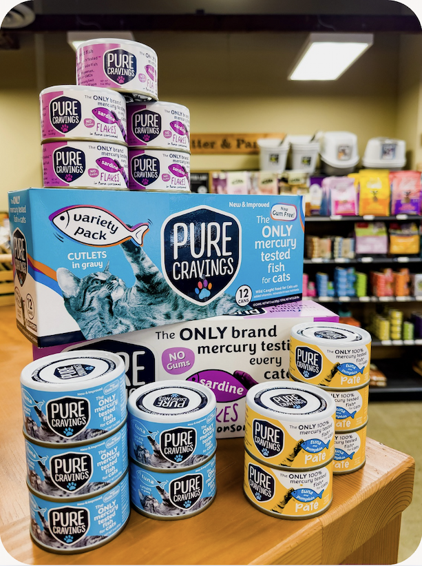 Photo of a variety of Pure Cravings canned cat food options displayed on a table inside a Mud Bay location.