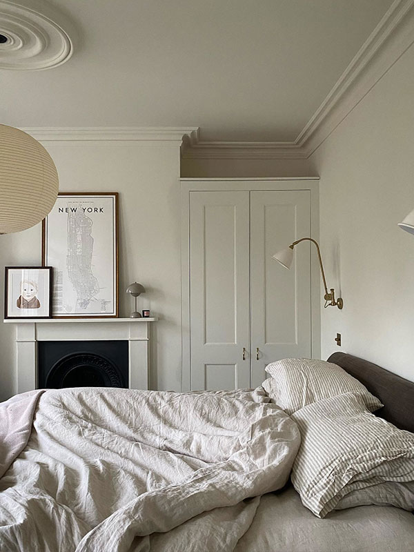 A softly lit bedroom with rumpled linen bedding, pale walls, and a framed New York map above a small fireplace. Calm and understated in tone.