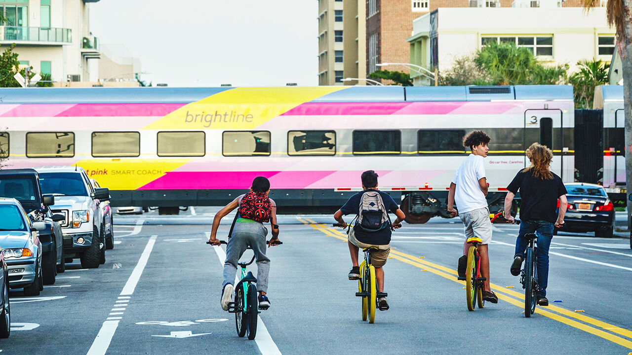 Teens biking near a crossing where a Brightline passenger train is passing.