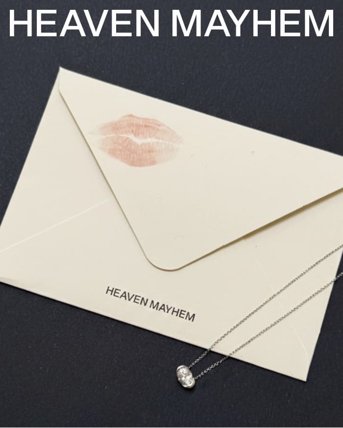 Heaven Mayhem cream envelope sealed with a soft pink lipstick mark, styled on a dark surface with the Poise Necklace in silver draped across the front.