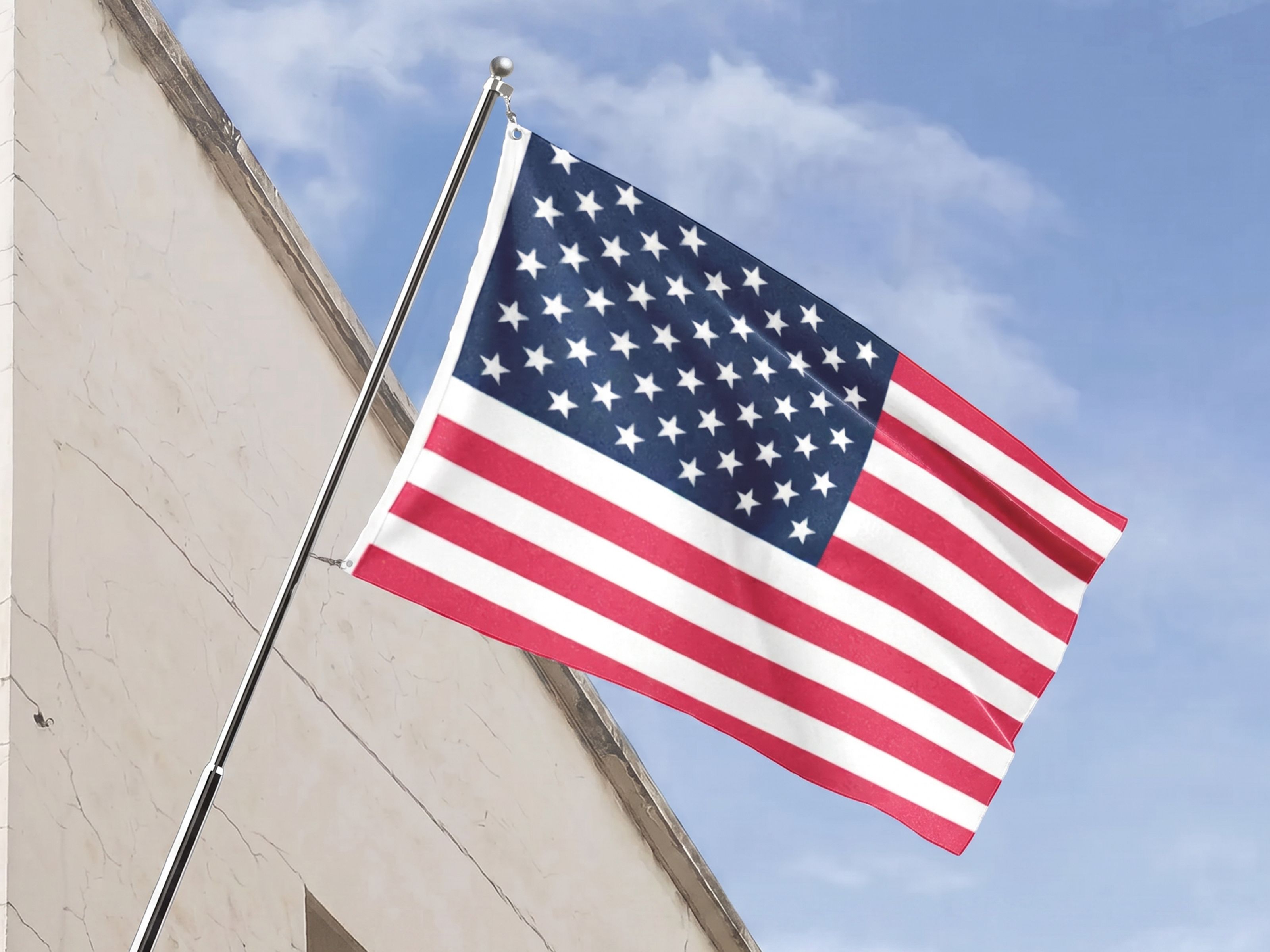 American flag mounted on an angled wall-mounted flagpole outside a building, waving against a cloudy blue sky. The flag’s red and white stripes and blue field with white stars create a patriotic display commonly used for Independence Day, national holidays, and civic celebrations.