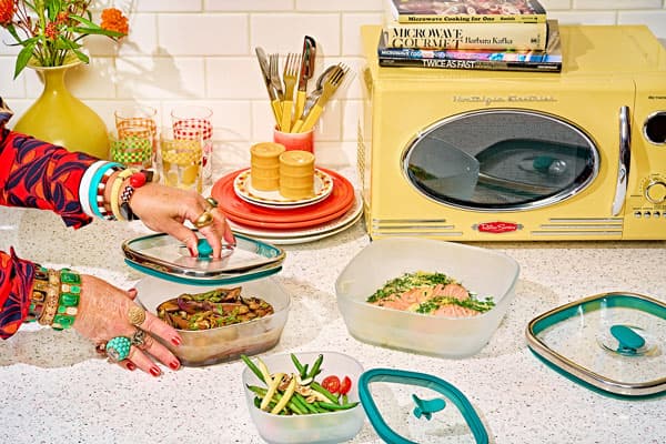 A vibrant, retro-inspired kitchen scene featuring a yellow vintage-style microwave, colorful glassware, and stacked plates. A person wearing bold jewelry is handling frosted Anyday square dishes filled with salmon, sautéed eggplant, and green bean salad, showcasing microwave meal prep. A stack of microwave cookbooks sits on top of the microwave.