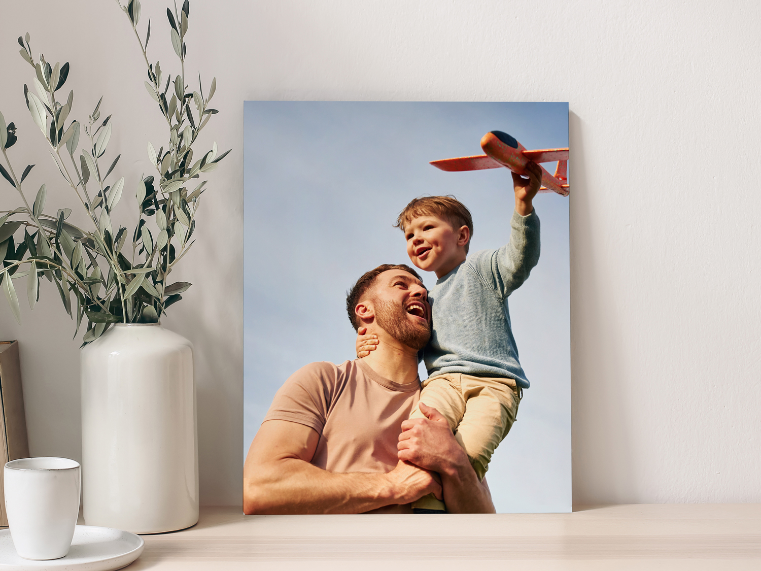 Foam board photo print displayed on a tabletop, featuring a smiling father holding his young son while the child raises a toy airplane against a clear sky background, styled beside a white vase with greenery.