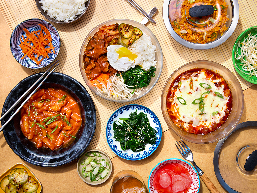Korean-inspired meal spread on a table with rice, side dishes, and mains served in Anyday microwave-safe dishes with Kale green lids.