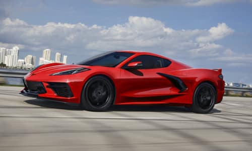 Side view of a 2024 Stingray Coupe in Torch Red driving down a roadway with a picturesque cityscape in the background.