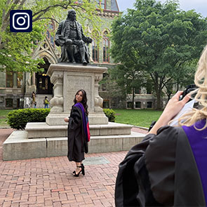 A student in a graduation robe takes a picture of another robed student in front of the Benjamin Franklin statue near College Hall.