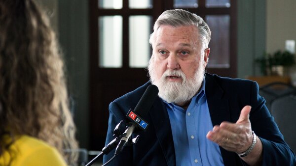 Doug Wilson is wearing a dark blue suit jacket and a light blue button-up shirt, talking at a mic with the NPR logo.