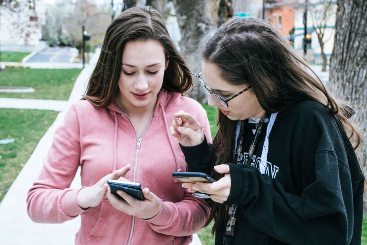 A young women in a pink hoodie touches the screen on her smartphones while second woman in black sweatshirt and glasses looks at the screen.
