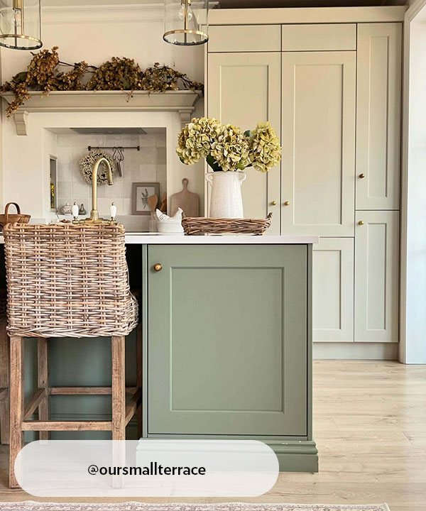 A soft, natural-toned kitchen with a mix of cream and sage-green cabinetry. Woven bar stools, brass details, dried foliage and flowers add country-inspired character, bringing warmth and subtle texture to the light, airy space.