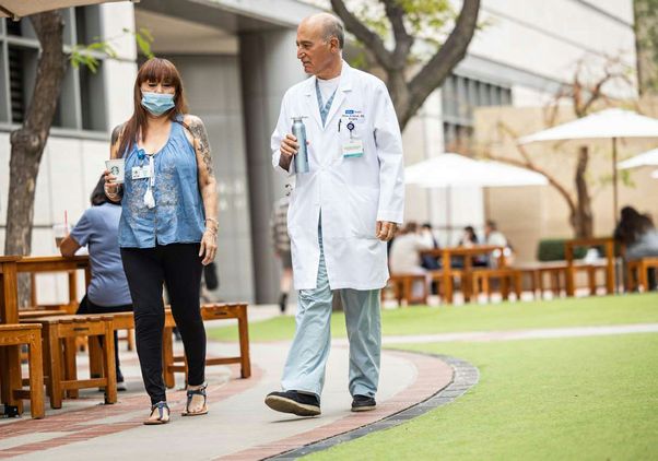 Dr. Abbas Ardehali, wearing a white lab coat over scrubs, walks outdoors with patient Patty Coles, who is wearing a protective mask.