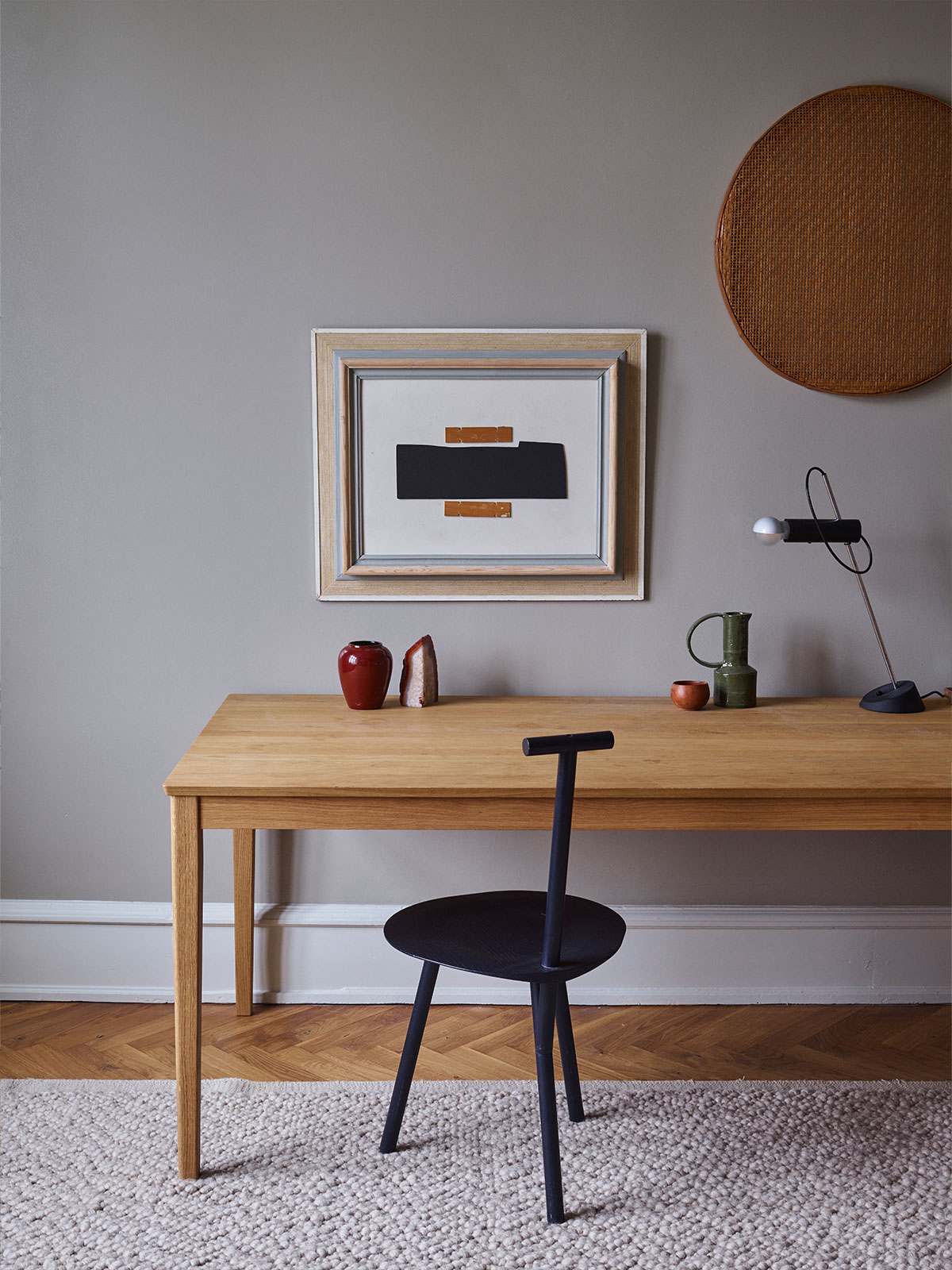 A minimalist study featuring a light oak desk against a muted grey wall. A black T-back chair sits on a chunky cream wool rug over herringbone parquet flooring. On the wall hangs a framed abstract artwork and a circular woven tray. Small ceramic vessels and a slender black desk lamp decorate the workspace.