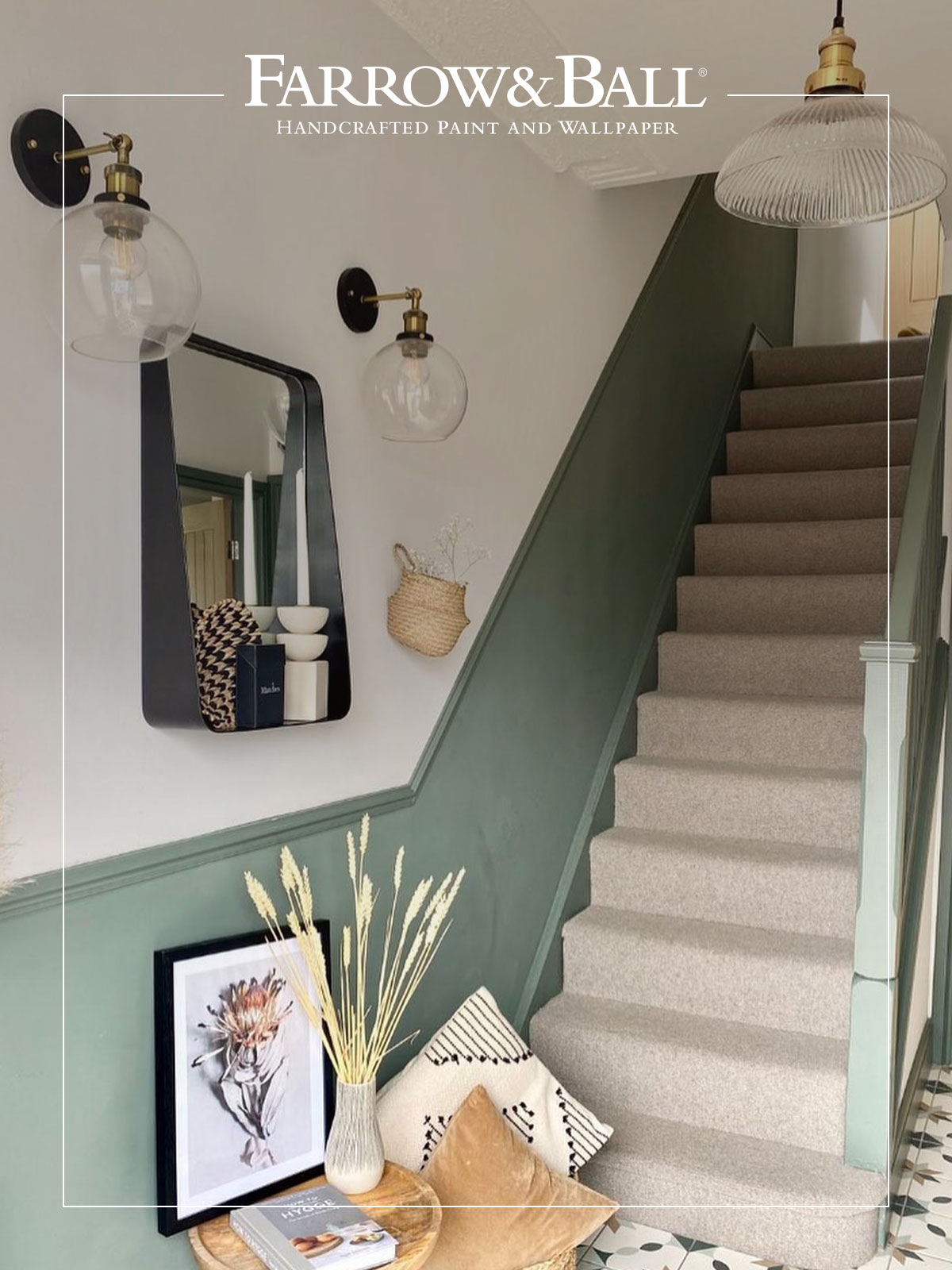 A bright hallway with a staircase painted in muted green and carpeted in beige. The wall features two glass globe lights with brass fittings, a black framed mirror, and a small woven basket. At the foot of the stairs, a wooden table holds a vase of dried grasses, a framed botanical print, and neutral cushions. The floor is patterned in soft green and white tiles. The Farrow & Ball logo appears at the top, highlighting handcrafted paint and wallpaper.