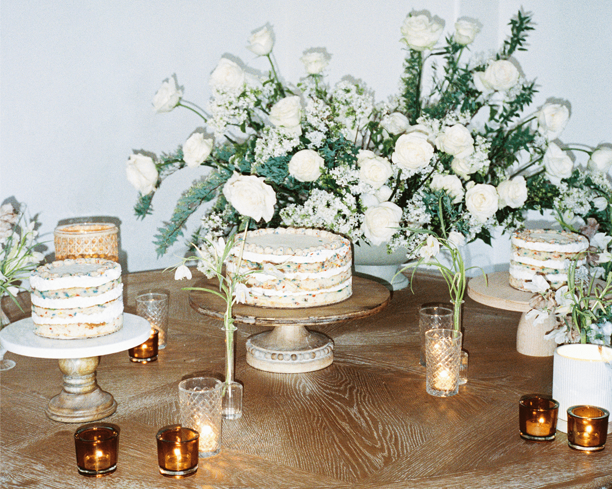 Three layer cakes displayed on stands on a wooden table, surrounded by lit candles and a large white floral arrangement in the background.