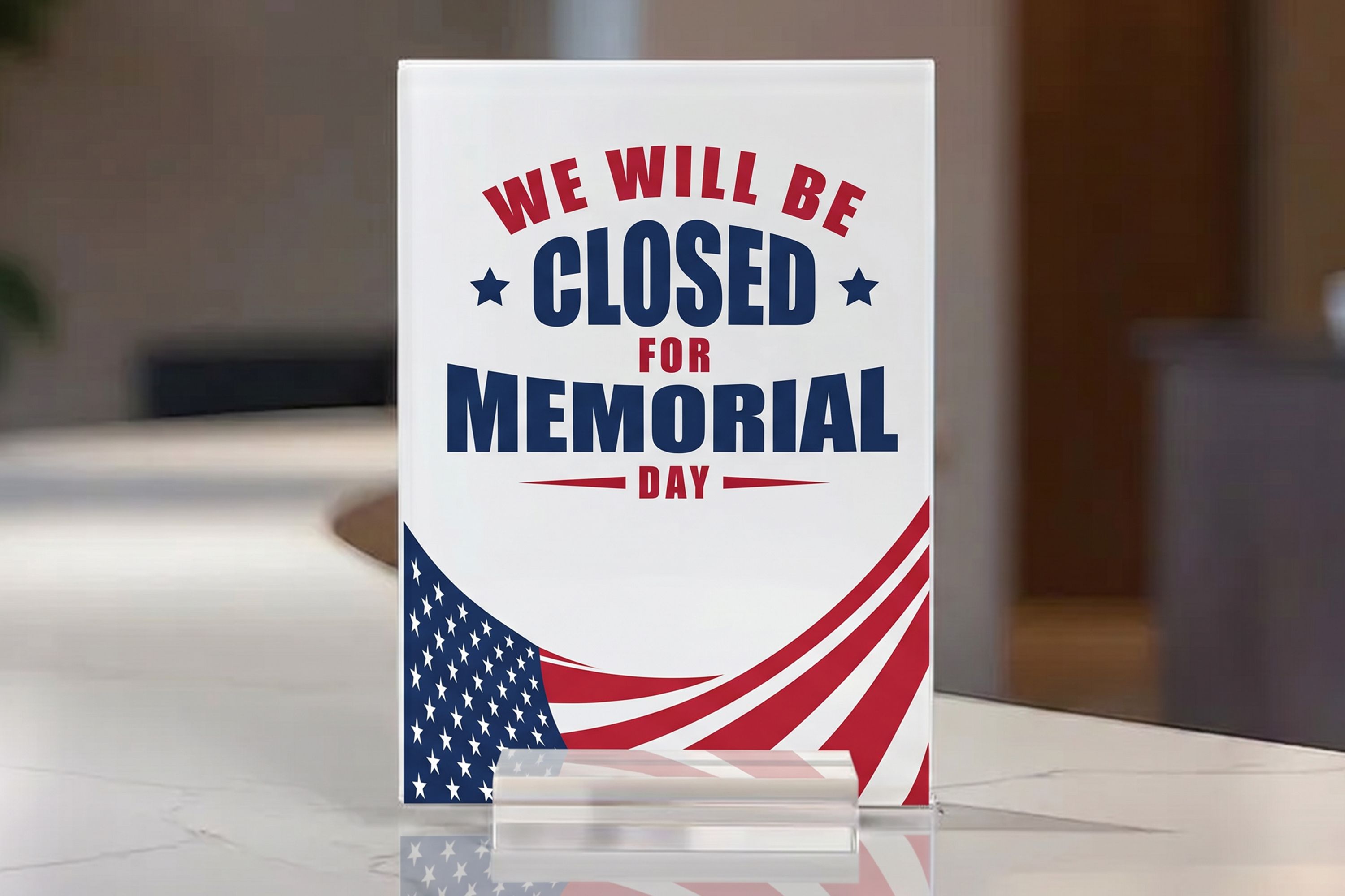 A acrylic table sign on a countertop displaying a red, white, and blue notice that says “We will be closed for Memorial Day,” with patriotic stars and flag-style stripes at the bottom.