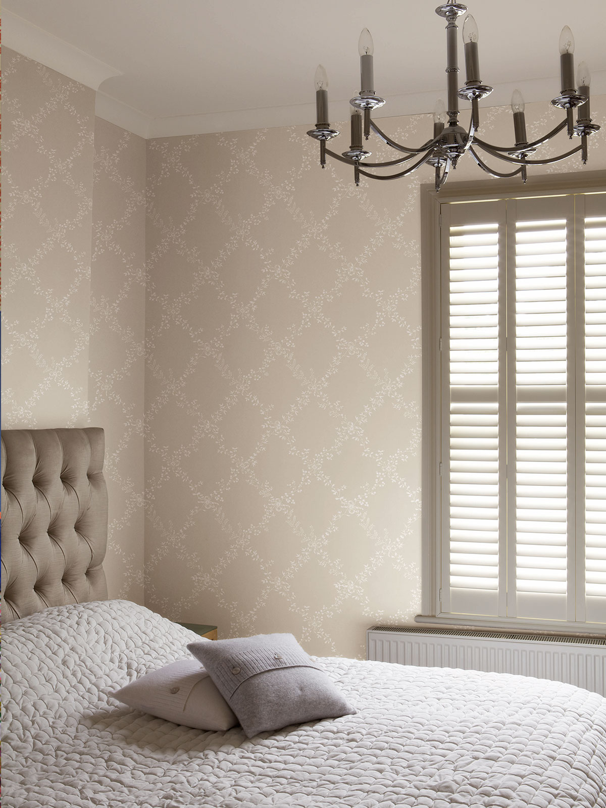 Soft neutral wallpaper and layered textures give this bedroom a calm, cocooning atmosphere. With gentle light coming through the window, an upholstered headboard and statement overhead light, the space feels elegant, restful and quietly luxurious.