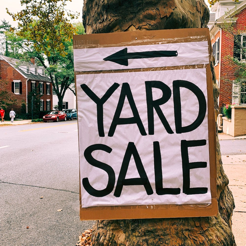 A big sign taped to a tree says YARD SALE in black letters. 