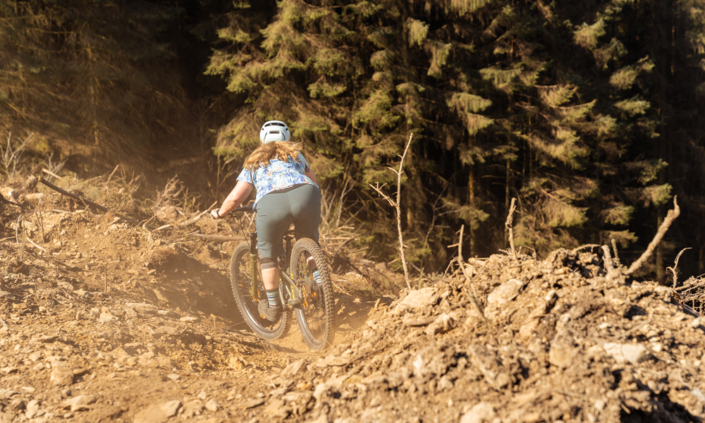 A view of a mountain biker from behind as she descends a steep, loose section of trail, with trees in the background.