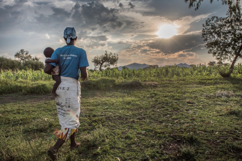 A woman carrying a baby walks through a green field toward the sunset.