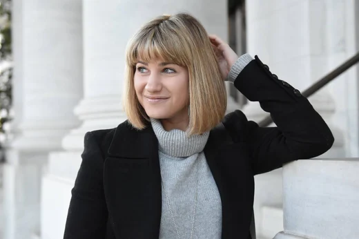 A professional headshot of a White woman with shoulder-length straight blonde hair and bangs wearing a light gray turtleneck sweater under a black coat. She has a subtle smile and looks slightly off-camera. One hand is raised, brushing her hair back. The background consists of white classical-style columns and a staircase, softly blurred.